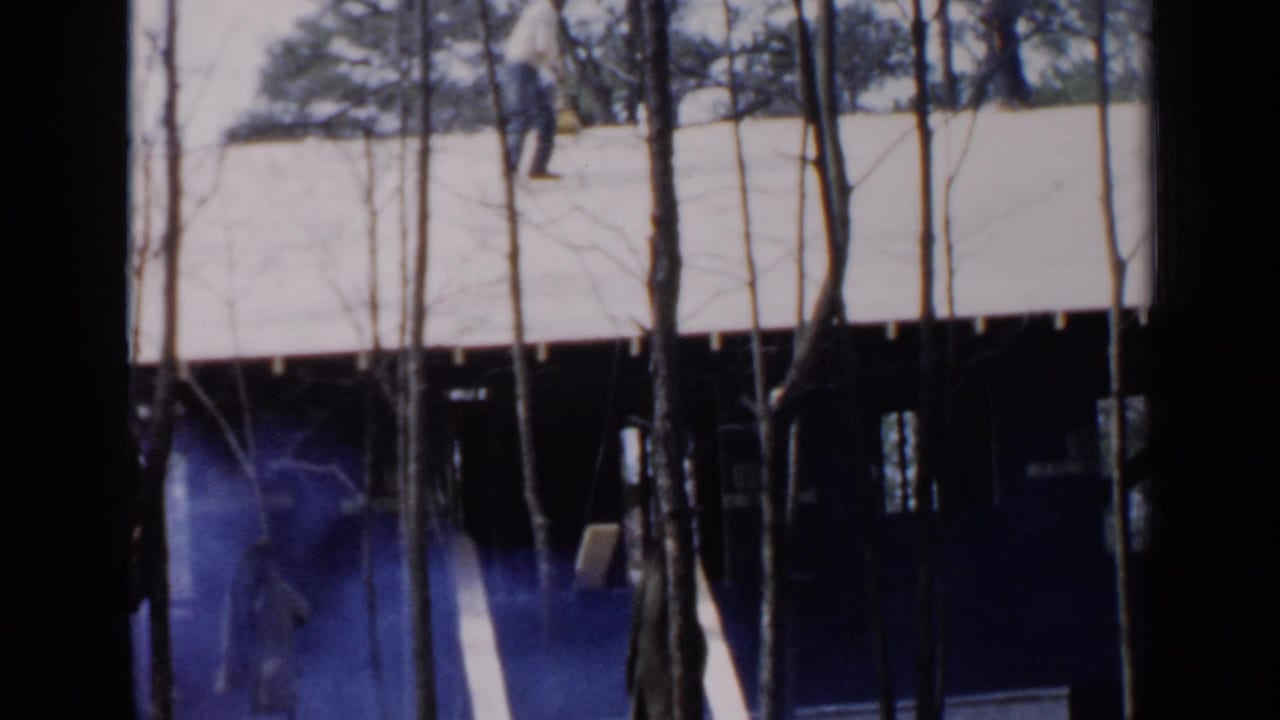 1957 Birmingham home-movie frame showing a house roof behind bare trees, with blue haze and a faint worker figure below.
