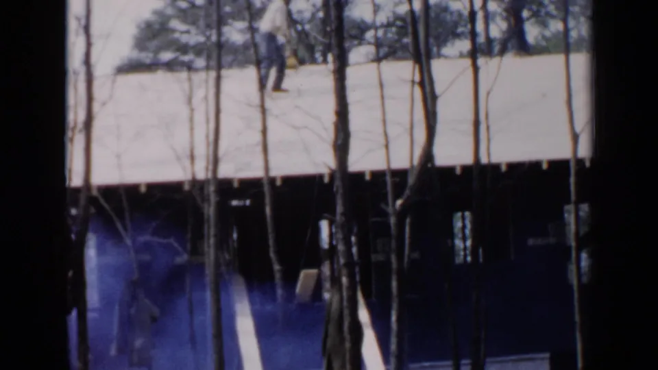 1957 Birmingham home-movie frame showing a house roof behind bare trees, with blue haze and a faint worker figure below.