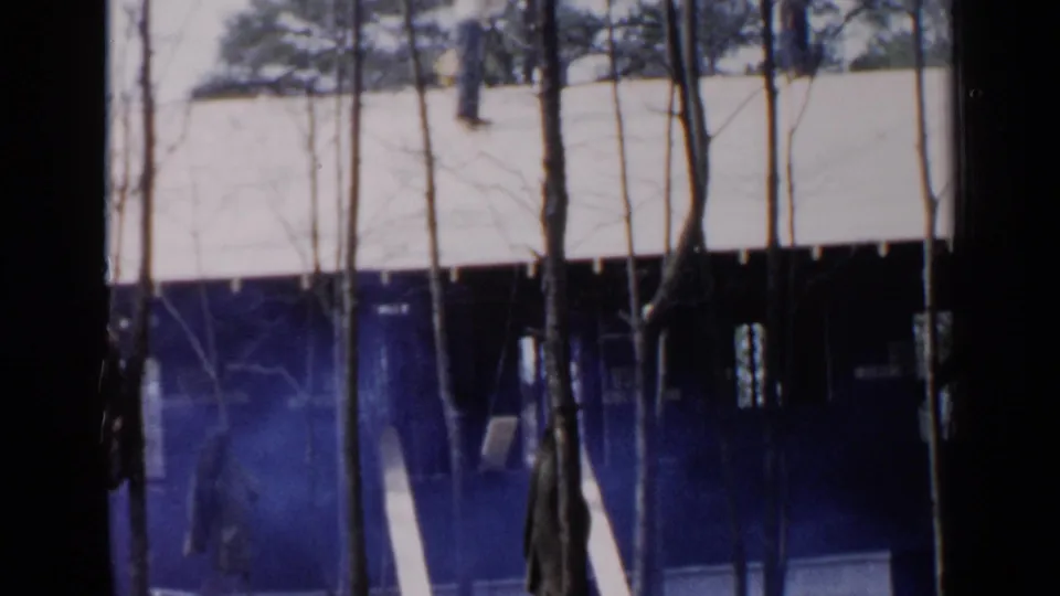 Blue-tinted 8mm frame of a Birmingham house roof partly blocked by trees, with a faint person visible near the lower level.