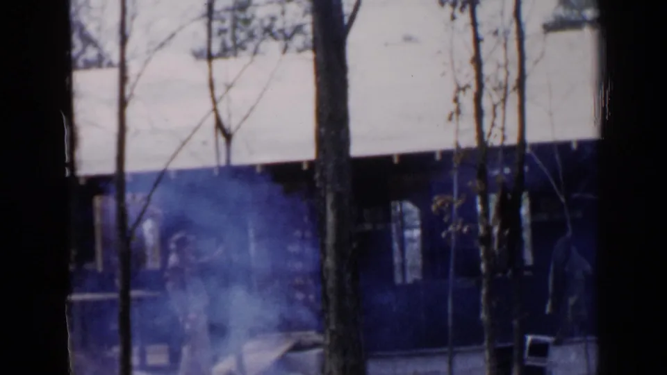 1957 home-movie frame showing bare trees in front of a Birmingham house, with blue haze and indistinct activity near the foundation.