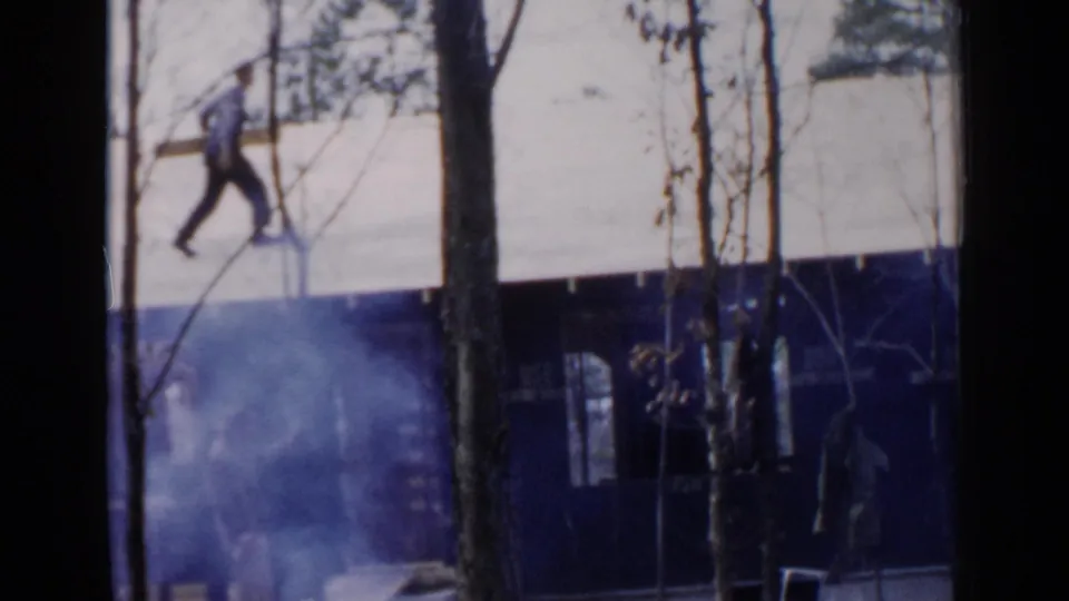 1957 Birmingham frame showing a man in blue pants carrying lumber on a house roof, with another distant figure near the lower level.