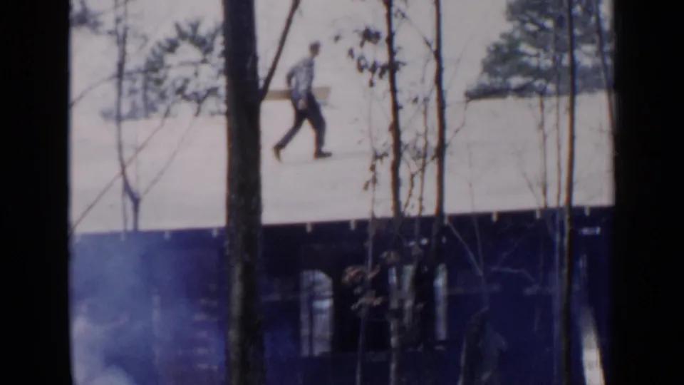 Distant rooftop figure carries a long board across snow, viewed through bare trees in Birmingham, Alabama metadata context.