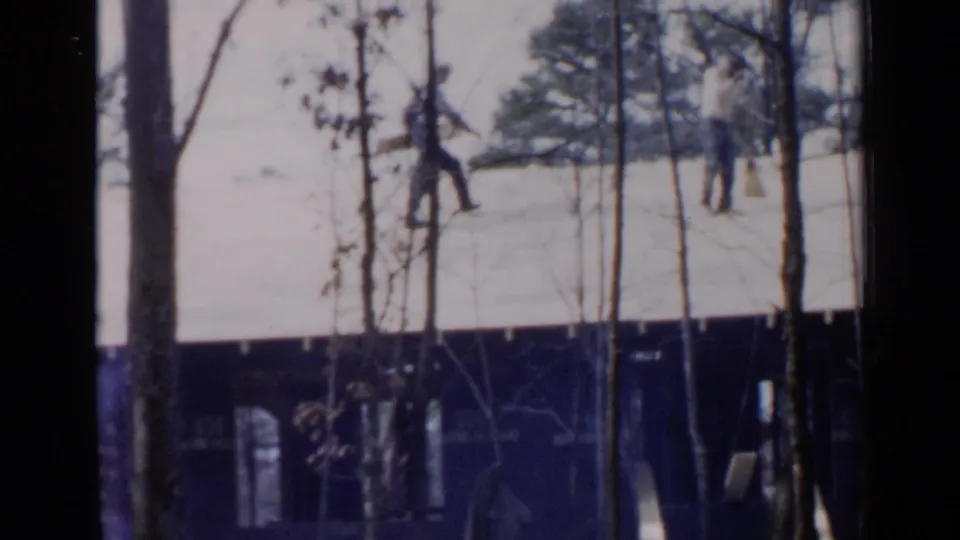 Two distant people stand or move on a snowy rooftop, with one near a long board and trees obscuring the view.