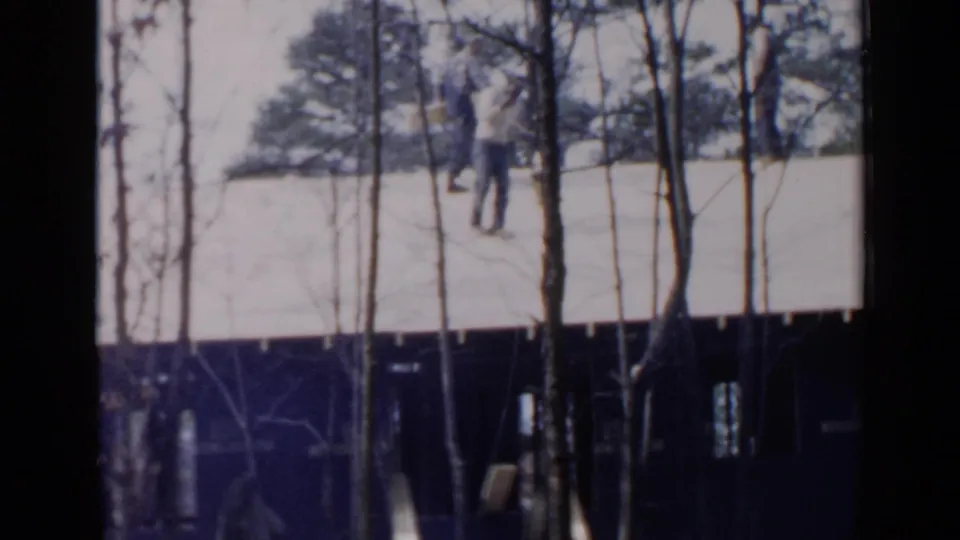 1957 Birmingham home-movie frame showing a roof worker on an unfinished house, partly obscured by bare trees and dark framing.