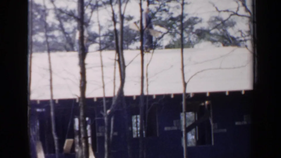 The roof and dark lower framing of a Birmingham house under construction appear behind leafless trees in this 1957 8mm frame.