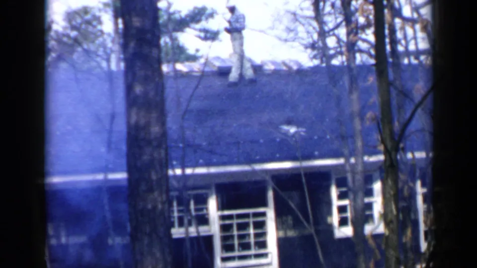 1957 Birmingham home-movie frame showing a man on a house roof with a broom or long tool, seen through bare trees.