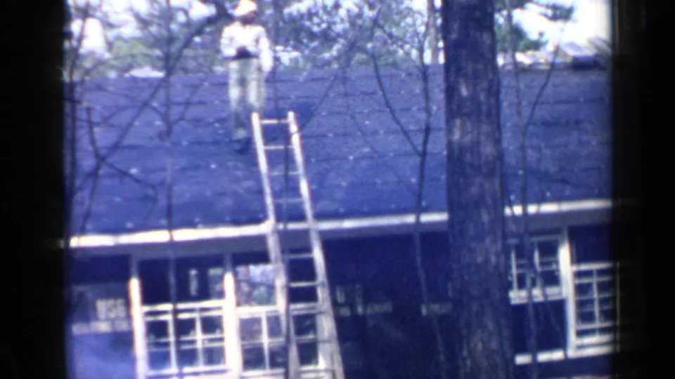 A man in a light shirt, pale pants, and hat stands on the roof near a ladder in the 1957 Birmingham residential scene.