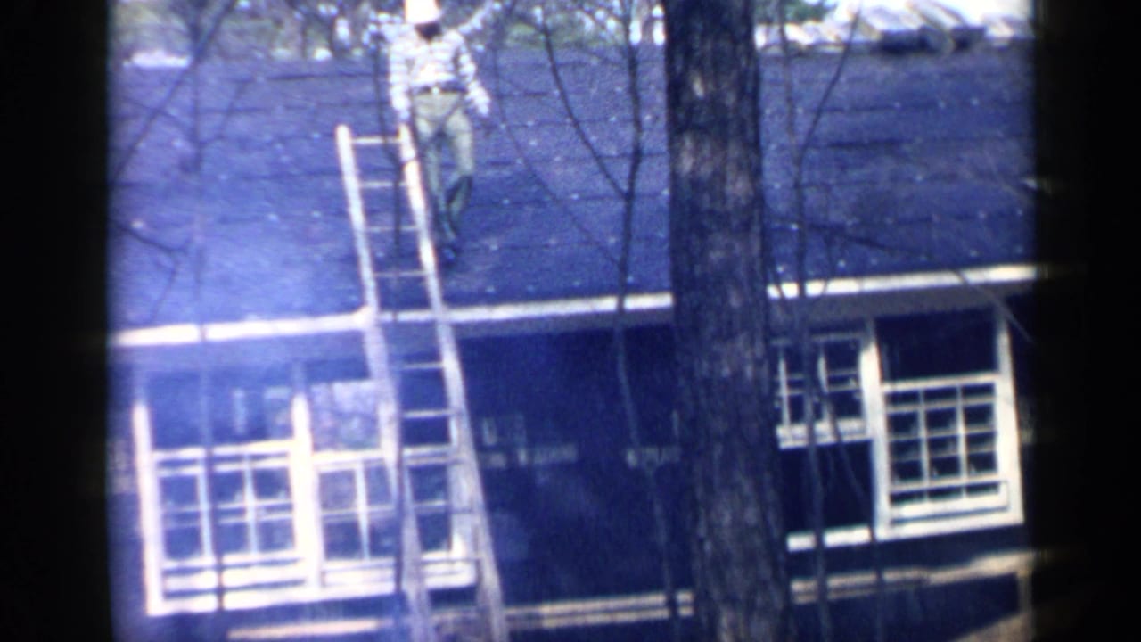In 1957 Birmingham home-movie footage, a man works near a wooden ladder on a dark shingled roof partly obscured by trees.