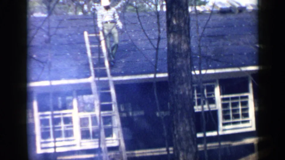 In 1957 Birmingham home-movie footage, a man works near a wooden ladder on a dark shingled roof partly obscured by trees.