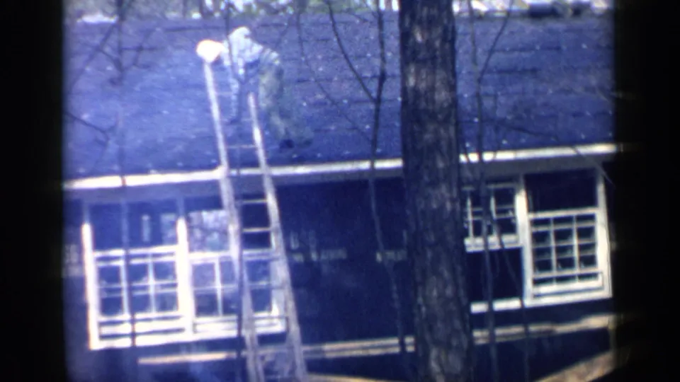 The man descends a wooden ladder from the shingled roof of a Birmingham house, with bare branches in the foreground.