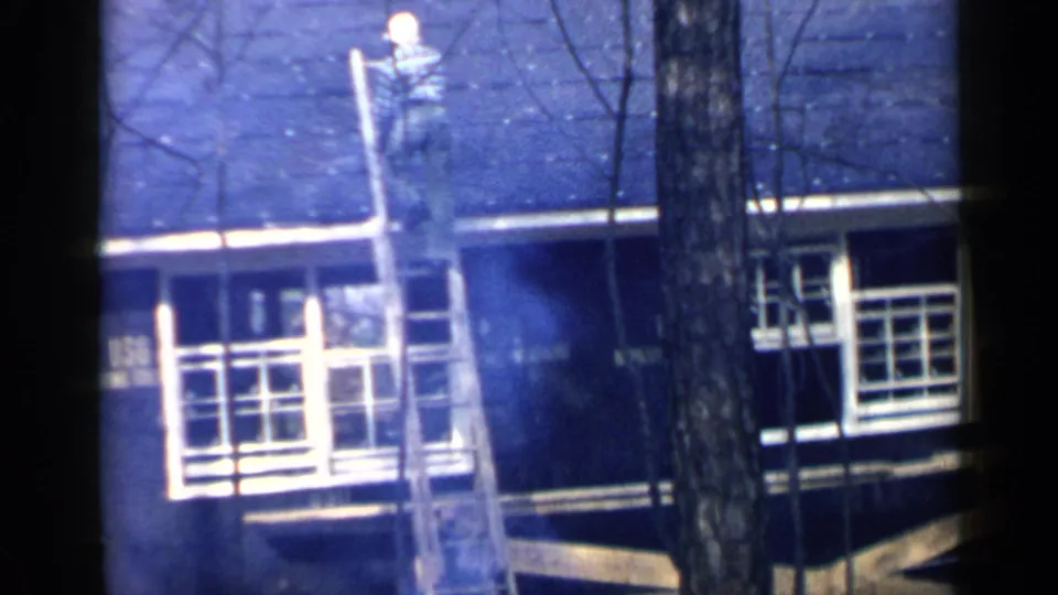A man in work clothes climbs down the ladder from the roof, framed by tree trunks and the white trim of the house.