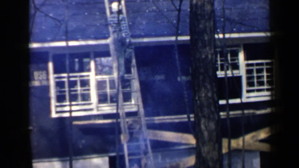 Birmingham, Alabama 1957 home-movie frame showing a dark house exterior, ladder, white-trimmed windows, and tree trunks.