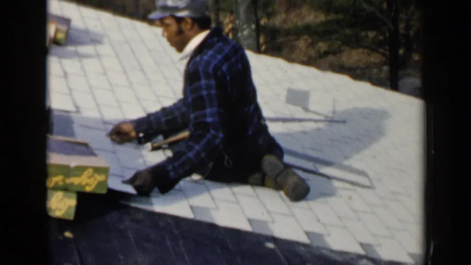 Birmingham, Alabama 1957 frame showing a man in a blue plaid shirt and cap kneeling on a roof while working with shingles.