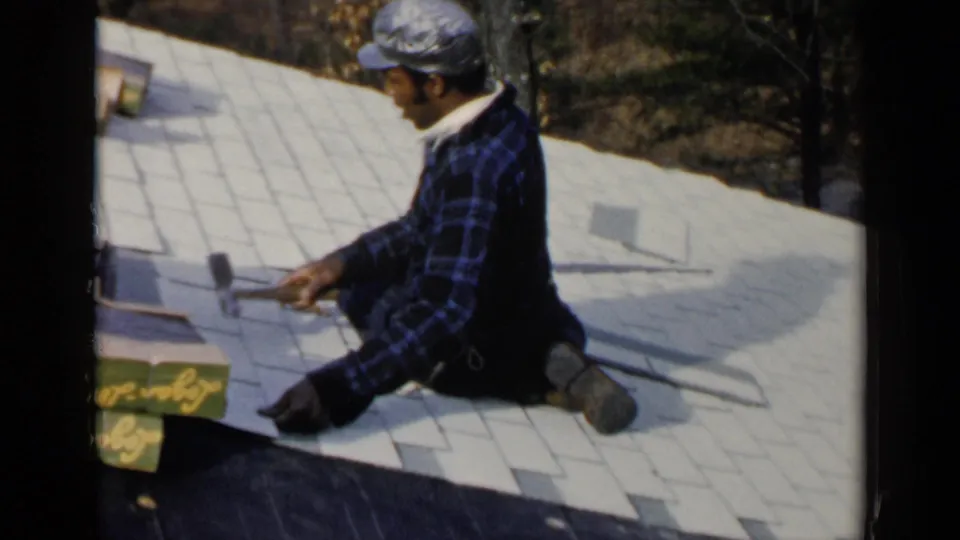 Birmingham, Alabama 1957 frame of a man kneeling on a shingled roof, holding a hand tool while installing or adjusting roofing material.