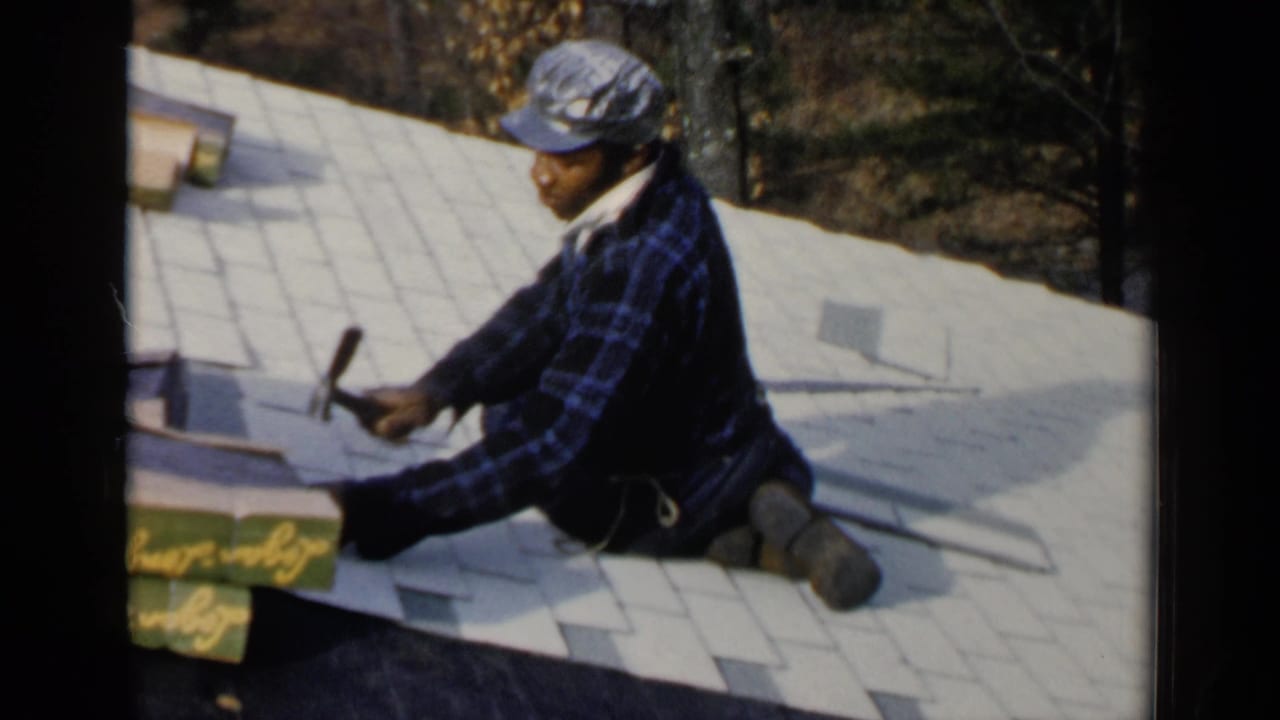 Roofer in plaid shirt kneels on a shingled roof while using a hammer.