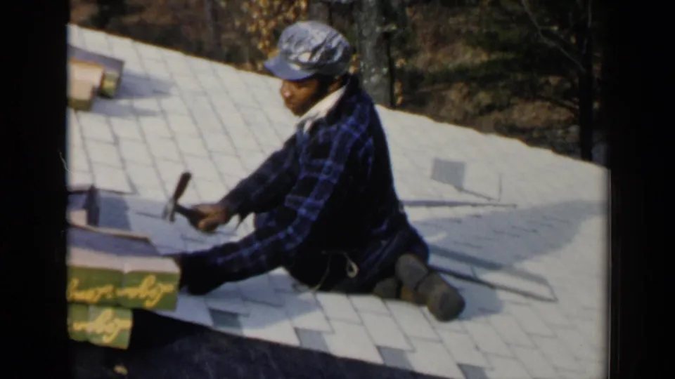 In Birmingham, Alabama, 1957, a roofer in a plaid shirt and cap kneels on a roof and hammers shingles into place.