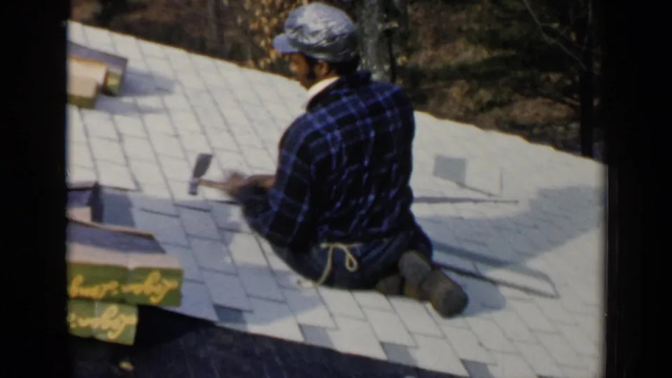 A roofer in a cap and plaid shirt sits on the shingled roof in Birmingham, Alabama, with tools and loose roofing material nearby.