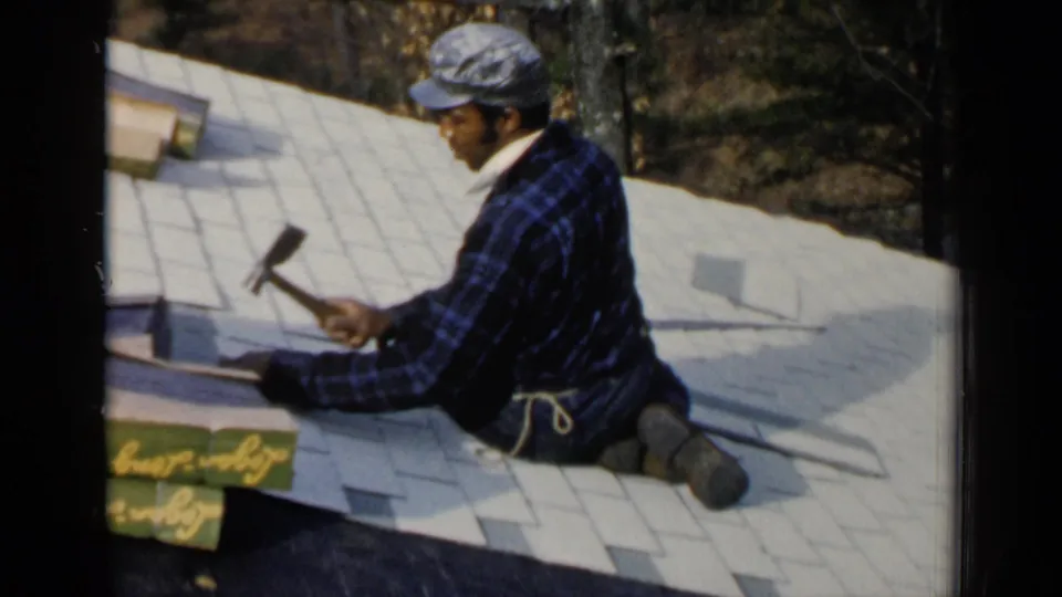 A 1957 Birmingham roofing scene shows a kneeling worker hammering shingles near visible roofing packages on a light-colored roof.