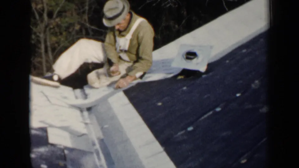 A second worker in a light hat and overalls appears on a Birmingham roof, working beside darker roofing material in 1957 home-movie footage.