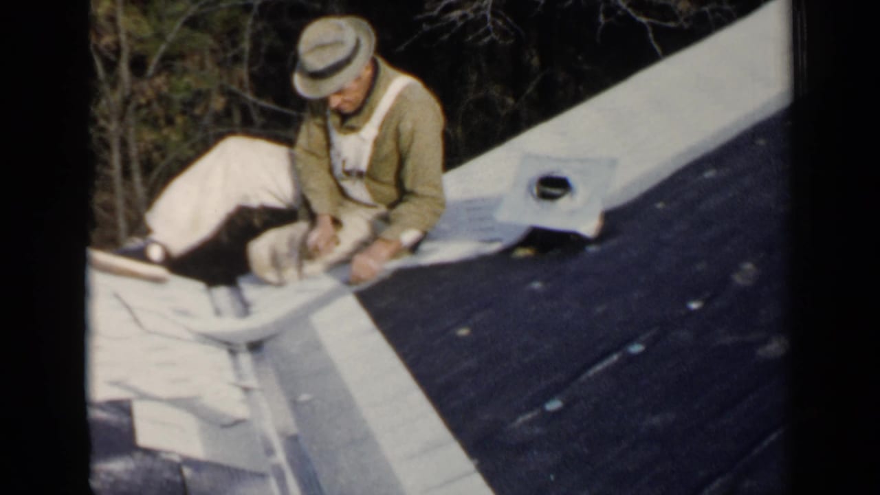 Man in hat and overalls kneeling on a roof beside pale flashing.