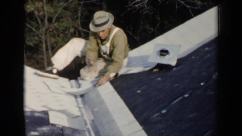 The man remains kneeling on the roof in Birmingham, Alabama, working near a pale roof edge or metal seam.