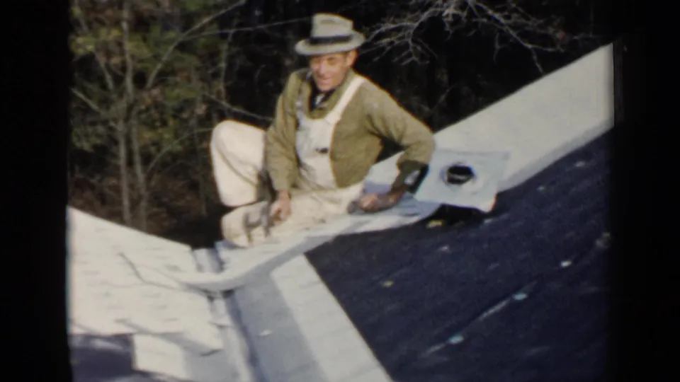 A man in overalls and a green jacket sits on the roof with one knee raised, framed by trees in 1957 Birmingham footage.