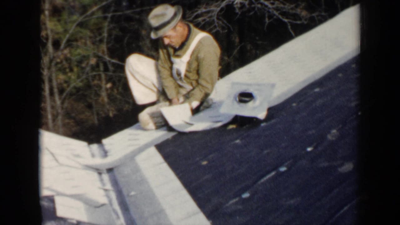 Man in a fedora kneels on a dark sloped roof with light roofing material nearby.