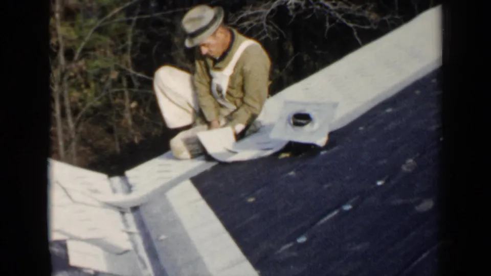 In 1957 Birmingham home-movie footage, a man in a fedora and overalls kneels on a dark roof beside light roofing material.