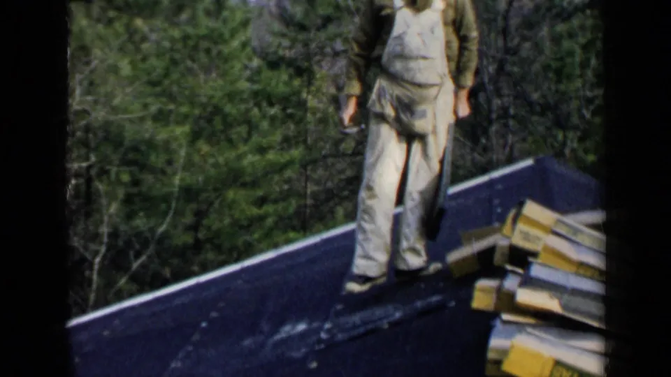 A man in light work clothes stands on the sloped roof near stacked boards or shingles, with trees in the background.