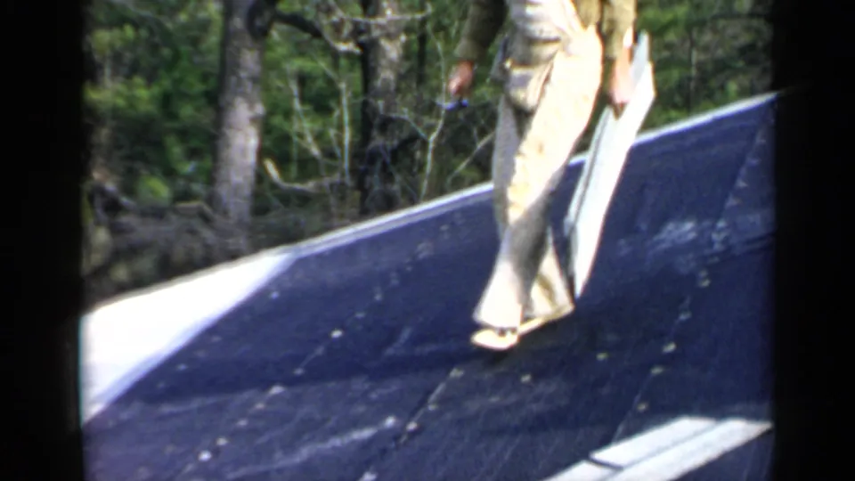 A man in overalls moves across the dark sloped roof while carrying light roofing material in the wooded residential setting.