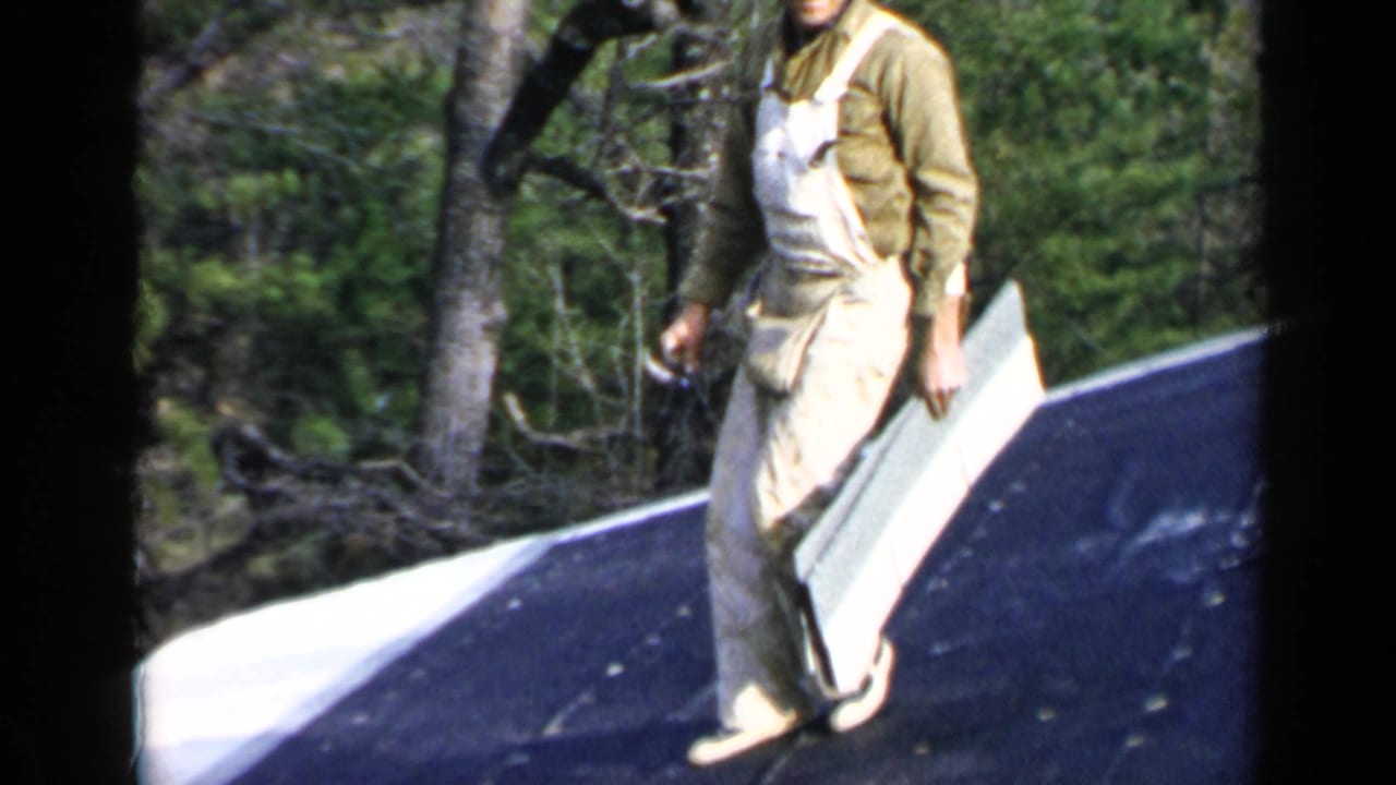 Worker in overalls carries a pale sheet on a sloped roof near trees.