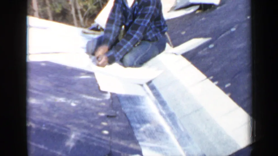 A worker in a plaid shirt and cap kneels on the sloped roof beside pale roofing material in the Birmingham roof-work scene.