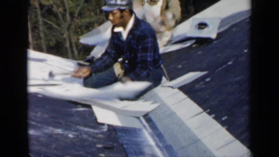 In the 1957 Birmingham home-movie clip, a worker in a plaid shirt and cap hammers pale material on a sloped roof.