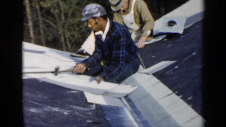 A plaid-shirted worker hammers roofing material while a second worker in overalls stands behind him on the sloped roof.