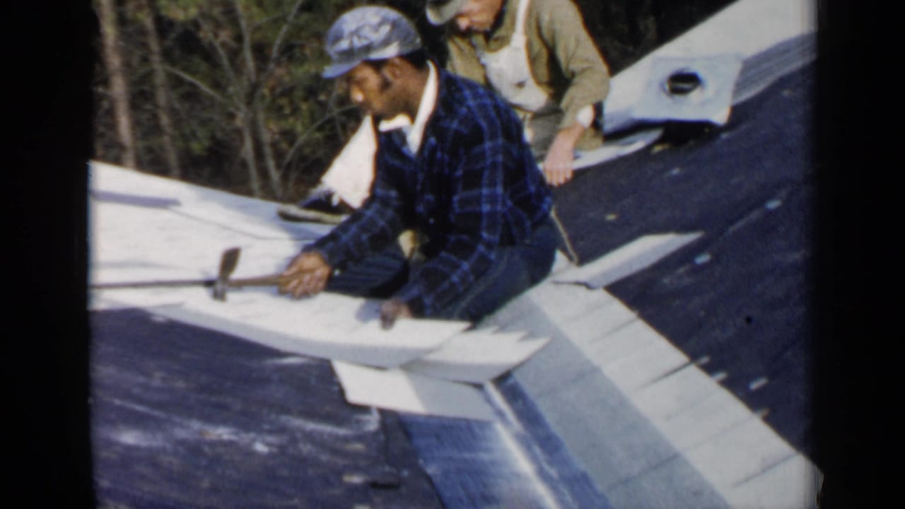 Man in blue plaid shirt kneels on a roof while holding a hammer near pale roofing material.