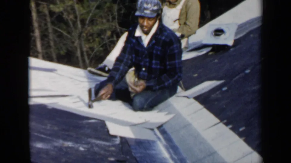 A roofer in a blue plaid shirt kneels on a Birmingham roof while another worker in overalls appears behind him near the shingles.