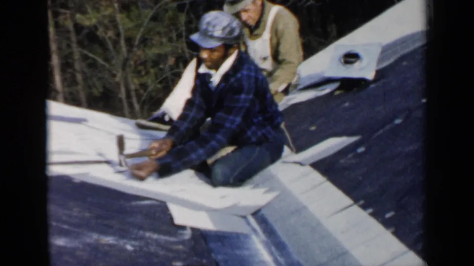 The 1957 home-movie frame shows a man in plaid leaning into roofing work with a hammer, with another worker behind him.
