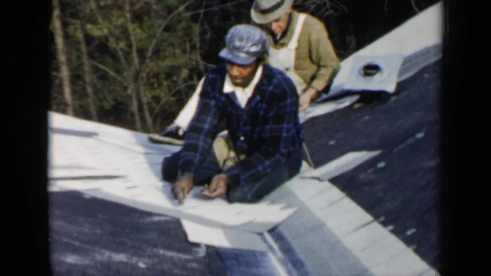 Two men work on a roof in Birmingham, Alabama, with the front worker kneeling near a hammer and pale roofing material.