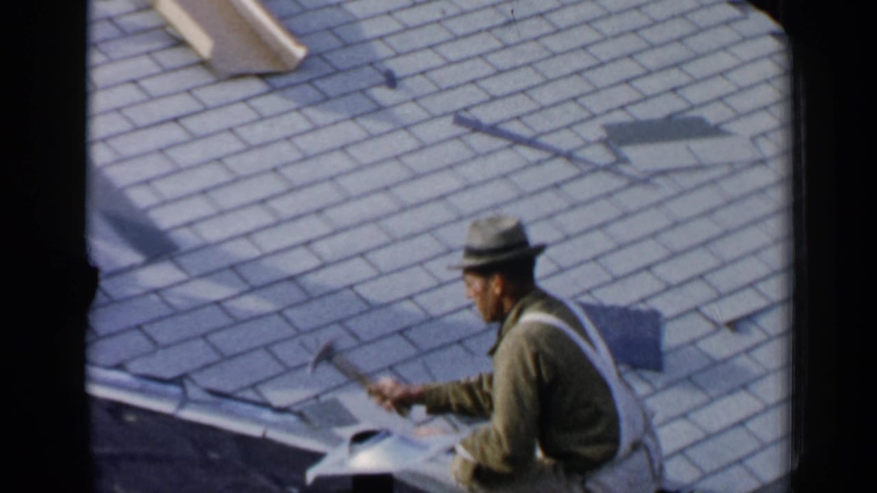 Man in work clothes sits on a shingled roof holding a tool near loose roofing material.