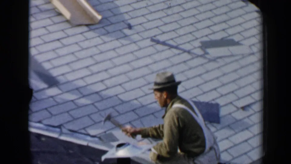 In 1957 Birmingham, Alabama, a man in a hat and work clothes sits on a residential roof while handling a tool beside asphalt shingles.