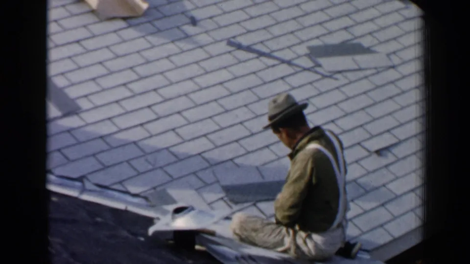 The worker remains seated on the Birmingham roof, turned partly away beside loose asphalt shingles and roofing tools.