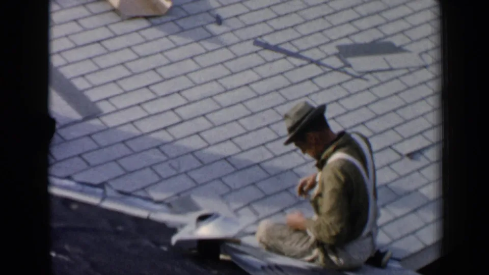 A man in work clothes leans over roofing material on a Birmingham residential roof, continuing shingle installation or repair.