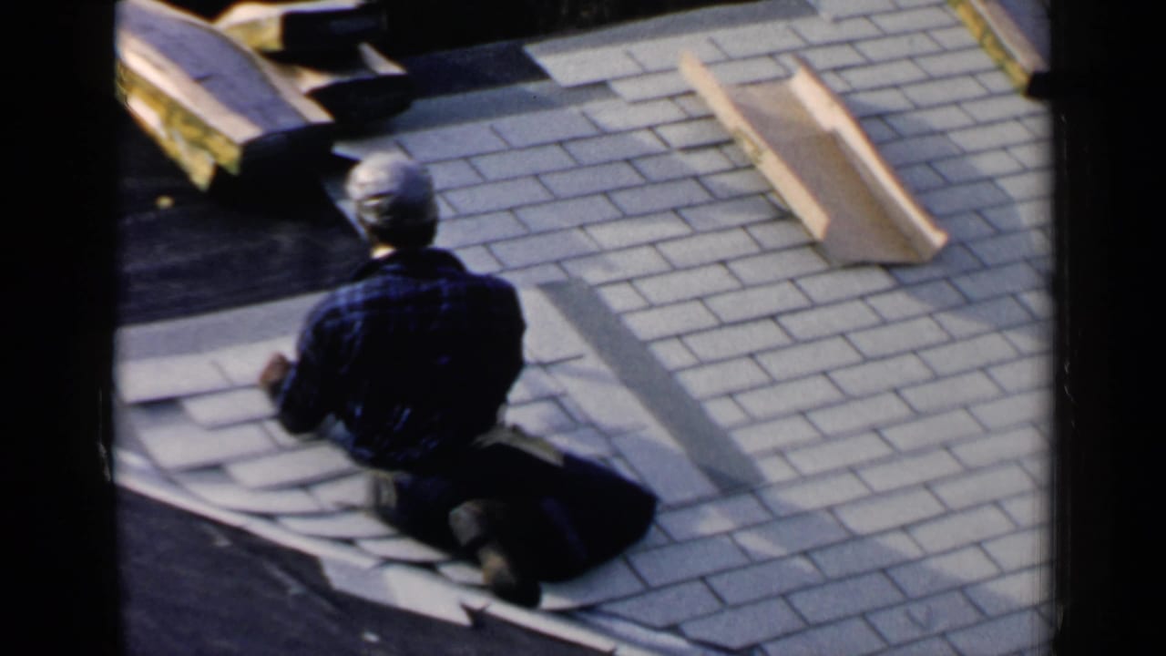 Man kneels on a roof while working with shingles beside loose roofing material.
