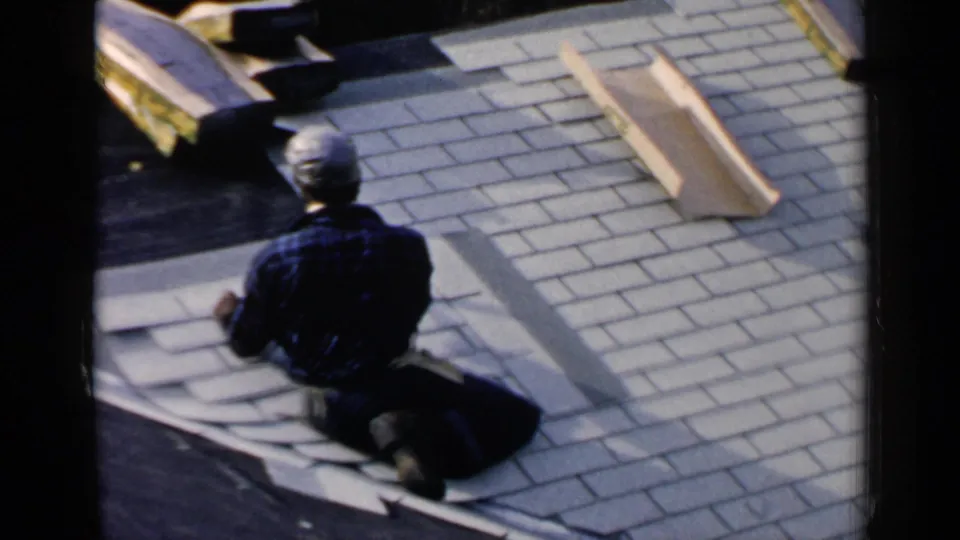 In 1957 Birmingham home-movie footage, a man in a plaid shirt kneels on a residential roof while placing shingles.