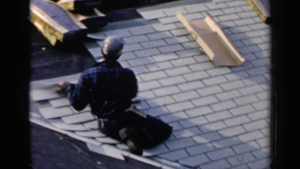 A man in a plaid shirt and cap continues shingle work on a residential roof in the 1957 Birmingham clip.