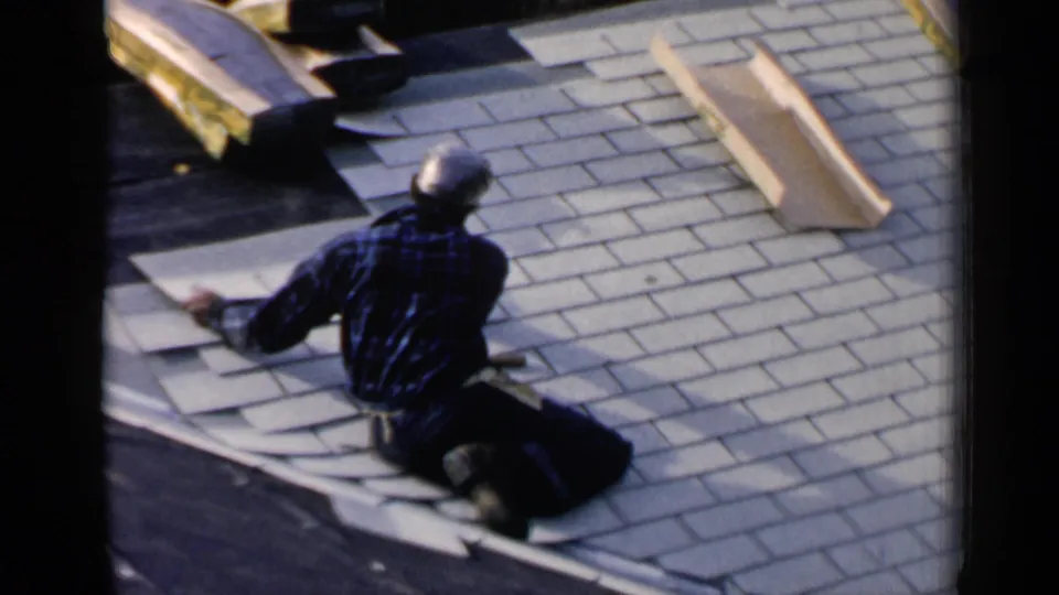 The home-movie frame shows the man reaching over roofing sheets as he works across the shingled roof surface.
