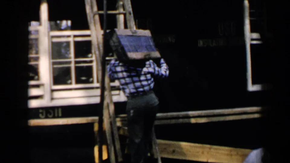 A man climbs a wooden ladder with roofing material near a house, with a USG Insulating Board sign visible in the background.