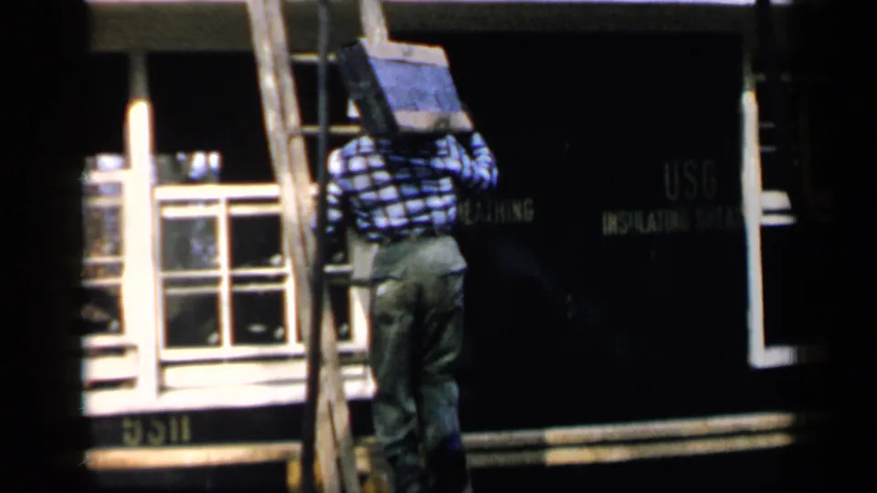 In 1957 Birmingham home-movie footage, a man in a plaid shirt climbs a wooden ladder with a dark panel near a house roof.