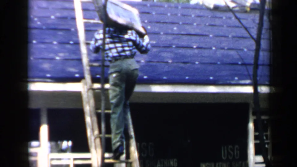 A rear view shows the plaid-shirted man balancing on the wooden ladder with the panel near the roofline.