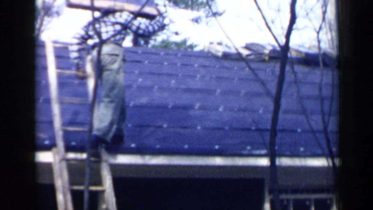 Man climbs a ladder onto a dark shingled roof carrying roofing material.
